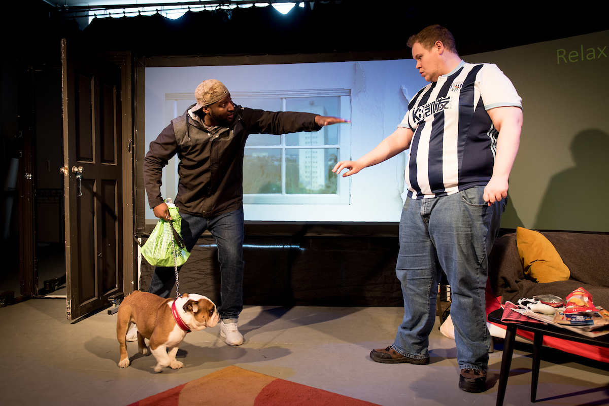 . The young man known as Bubble is in his flat, he wears a West Bromwich Albion football shirt, he opens the door and gestures with his hand. He is trying to stop the dog entering his flat