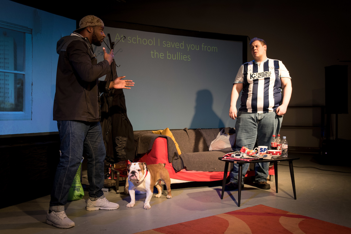  The man has a cap on his head. The peak of the cap is on the back of his head. The man gestures with his hands. He is communicating the character of Troy and uses urban sign language.
