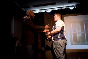 A man has entered the flat, the character is Troy. The man has a cap on his head. The peak of the cap is on the back of his head. The man gestures with his hands. He is communicating the character of Troy and uses urban sign language.
