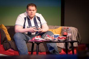 The young man looks like he does not do exercise. He is big and he wears a West Bromwich Albion football shirt, he sits on the sofa holding a game controller; he is surrounded by empty Crisp packets, bottles of Coke, Dr Pepper and old cups of tea. The character is known as Bubble