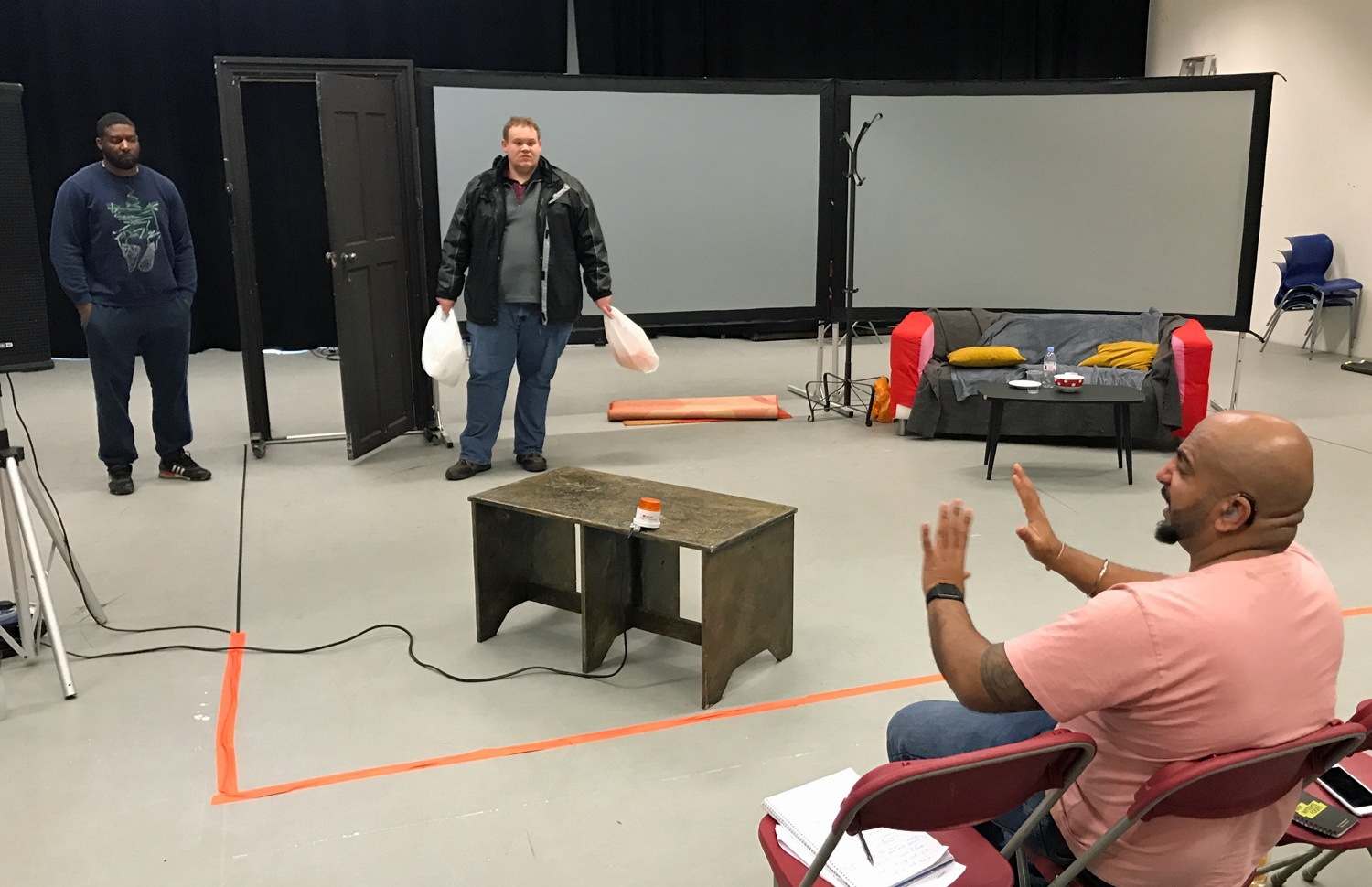 Man on right holds up hands He is directing two men in a room, with two screens, the man in the far left has his hands in his pockets, the man on his right clutches two full shopping bags