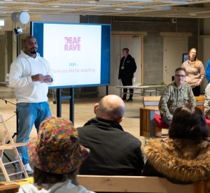 Two men sit with slide show, watched by an audience